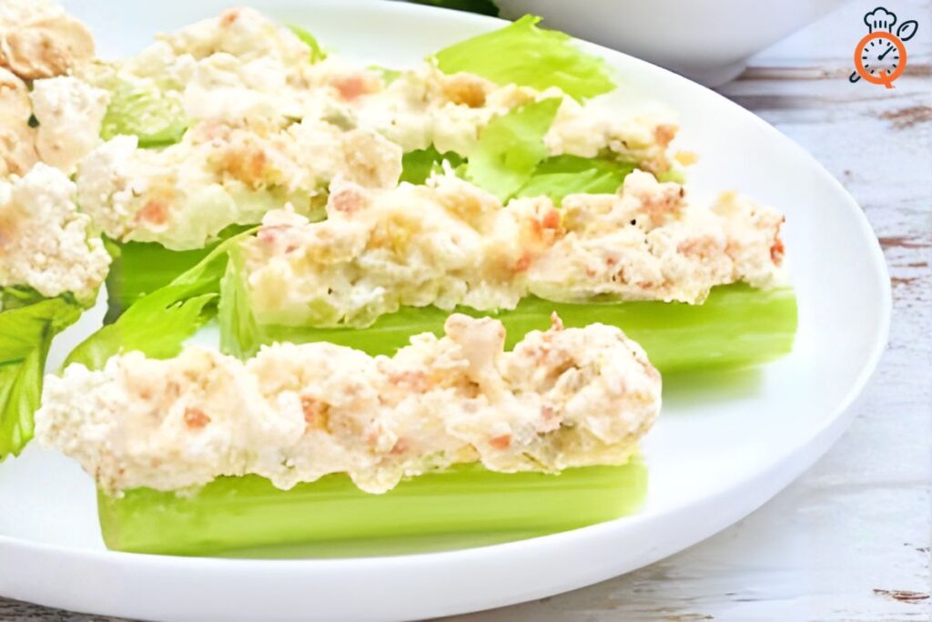 A close-up view of four bright green celery sticks on a white plate, generously loaded with creamy, savory stuffed celery fillings (possibly pimento cheese, bacon, or a tuna/chicken salad mixture), highlighting a delicious and easy appetizer.